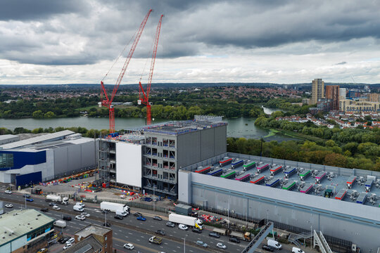 Aerial view of a large data center construction site featuring two tall red cranes, industrial buildings with rooftop cooling units, and busy traffic in the United Kingdom.