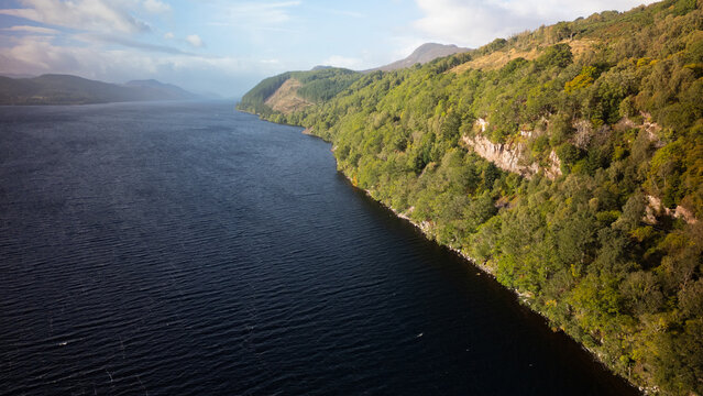 Aerial view of Loch Ness with its deep blue waters bordered by lush green forested hills and rugged cliffs in Highland Council, Scotland, United Kingdom.
