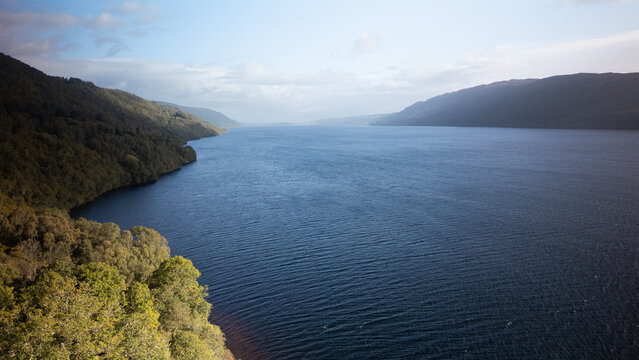 Aerial view of Loch Ness with its deep blue waters surrounded by lush green hills and dense forests under a bright sky in Highland Council, Scotland, United Kingdom.