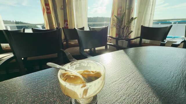 A delicious bowl of ice cream sits on a dark table inside a sunny cruise lounge. Passengers enjoy a peaceful, delightful moment taking in views of Lake Ladoga.