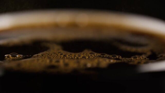 Freshly brewed coffee bubbles and foam shift across a dark surface in an extreme close-up cup view. Coffee advertising, beverage packaging, and caf promotion.