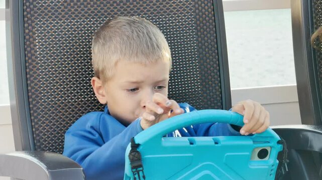A focused young boy holds a blue tablet while sitting on a cruise ship deck. He feels excited and cheerful during a smooth voyage across Lake Ladoga.