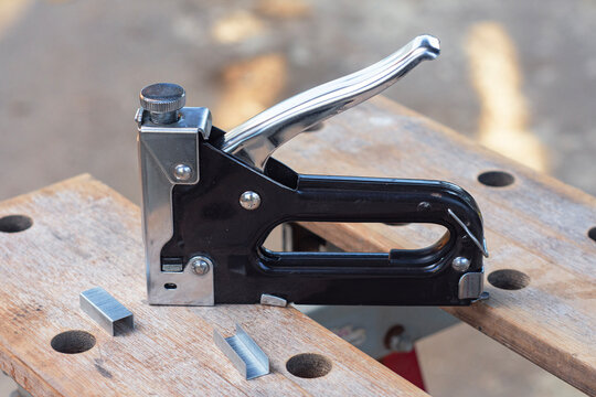 Manual staple gun with metal staples on wooden workbench close up. Upholstery tool, carpentry equipment or DIY construction hardware concept.