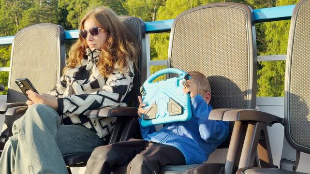 A mother and her young boy relax on deck chairs while checking their handheld devices on a cruise ship. They feel calm and peaceful sailing past lush green shores on Lake Ladoga.