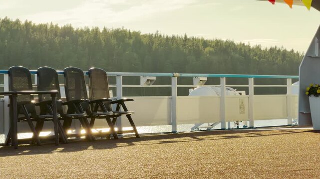 Empty chairs stand along a sunlit ship deck overlooking a wooded shore. They offer a calm and peaceful view on Lake Ladoga. The quiet atmosphere feels serene and relaxing.