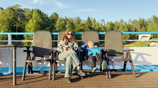A mother and her young boy sit on deck chairs aboard a cruise ship while the boy plays on a tablet. They feel peaceful and happy sailing past green trees on Lake Ladoga.