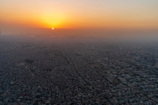 Aerial view of the sprawling urban landscape at sunset, with dense residential buildings stretching to the horizon under a golden hazy sky in Karachi, Sindh, Pakistan.