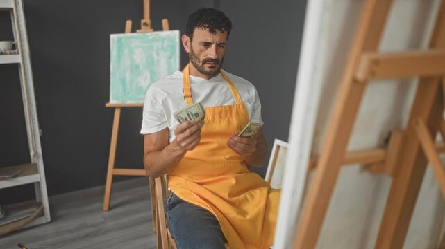 Seated man painter counting dollars with hands, wearing orange apron beside easel in studio; concern creative struggle.