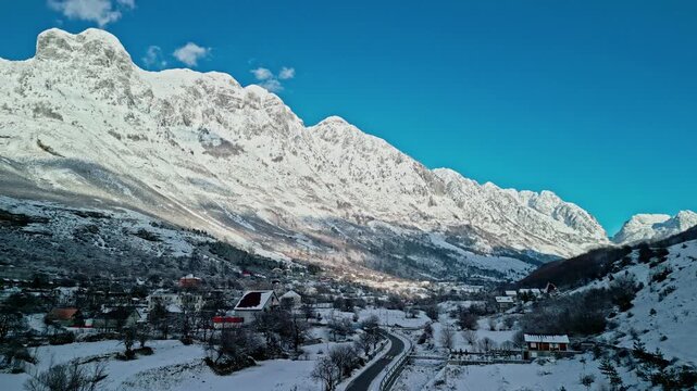 Albania, Boge 11 01 2026: Still 4K drone video of snow-covered Boge showing a car on the road, frozen Prroni i Thate riverbed, and sunlit Albanian Alps under a clear blue sky.