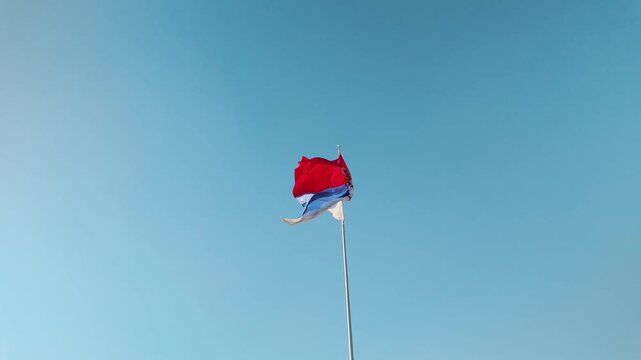 Serbian national flag waves in the wind against a clear blue sky. Symbolic footage represents Serbia, national identity, patriotism and state culture.