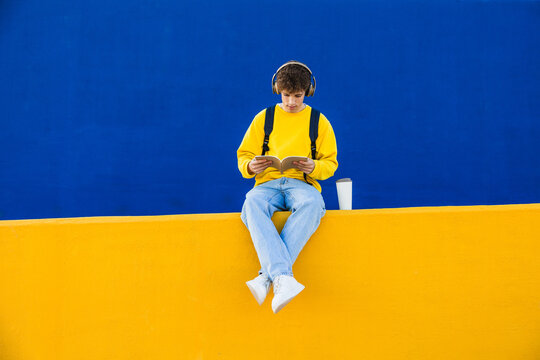 College student reading a book and listening to music with headphones in front of blue and yellow wall