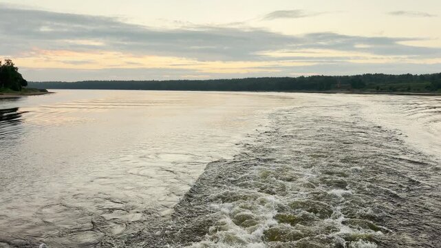 A boat wake leaves a foaming trail on the wide river. A calm and peaceful feeling fills the air during this serene evening journey.