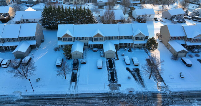 Aerial view of a suburban street featuring rows of townhouses covered in fresh snow, with snow-covered driveways and cars, under a clear winter sky in Manheim, Pennsylvania, United States.