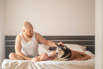 Man in tank top strokes head of husky resting under blanket on bed, tenderness that people rarely show openly, moment building real trust. Wide shot. Concept of tenderness between man and his dog © iwavephoto
