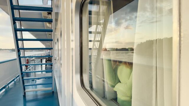 A woman looks out a cruise ship cabin window as it travels along a calm river. She enjoys a warm drink in a cozy moment. The peaceful landscape brings feelings of deep comfort and joy.