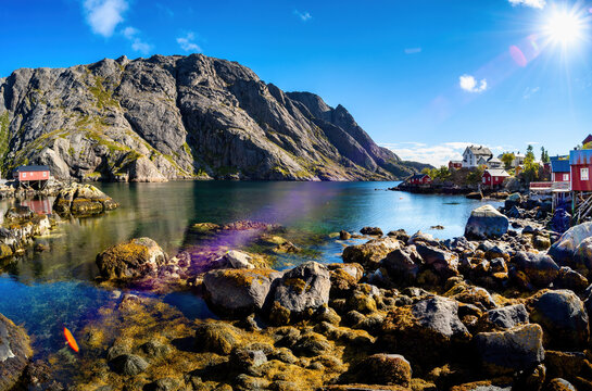 Traditional red and white wooden houses stand along the rocky coast of Nusfjord, surrounded by clear water and massive rocky mountains.