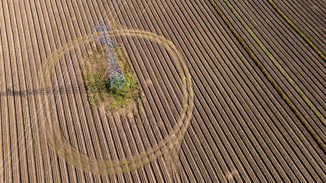 Aerial view of a high-voltage electricity pylon standing in the center of a large plowed agricultural field with circular tracks and parallel furrows in Poland.