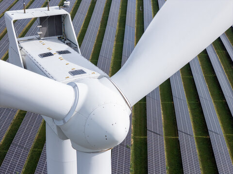 Aerial view of a large white wind turbine standing in front of rows of solar panels in a renewable energy farm in Poland.