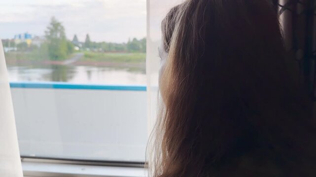 A woman gazes out a cabin window on a cruise ship. She watches the green shore pass along the calm river. The quiet view fills her with peaceful serenity and joy.