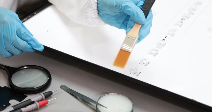 Forensic technician in blue gloves dusts fingerprint powder onto a document with a brush beside a magnifying glass and evidence tools.