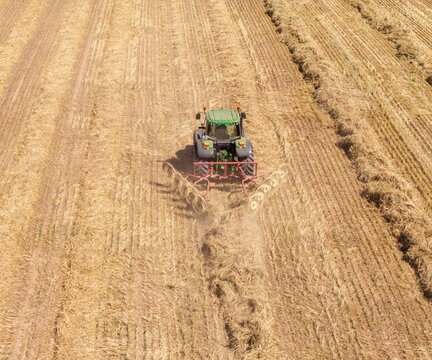 Aerial view of a green tractor pulling a hay rake through a dry, harvested field in Philadelphia, Western Cape, South Africa.