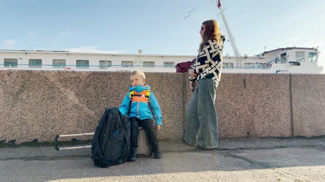 A boy sits on luggage while a woman stands near a docked cruise ship in Saint Petersburg. They wait happily along the Neva River embankment. The sunny morning brings calm excitement and joy.