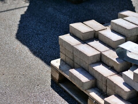 Stacked gray concrete paving bricks on a wooden pallet outdoors in bright sunlight with deep diagonal shadows.