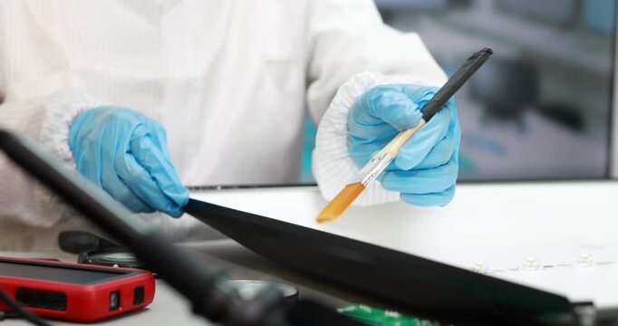 Close-up of a technician wearing blue gloves using a brush and tablet while repairing an electronic device on a workbench.