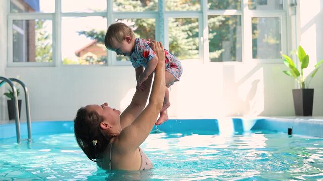 Mother lifting cute 11 months old baby girl up in air inside sunny pool