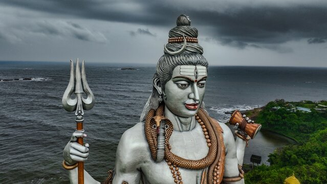 Aerial view of the massive Shiva statue at Murudeshwar Temple, holding a trident and damaru, set against the Arabian Sea and a dramatic cloudy sky in Murdeshwar, Karnataka, India.
