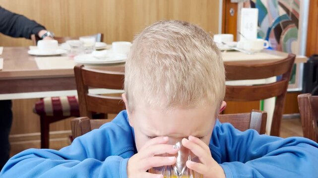 A young boy drinks juice inside a cruise ship dining room on Lake Ladoga. He holds his glass carefully with both hands. The warm breakfast brings feelings of sweet comfort and quiet joy.