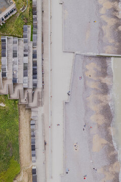 Veules-les-Roses, France - 18 June 2025: Aerial view of the beach promenade with rows of small wooden beach huts, stone walls, and people walking along the pebbled shore.