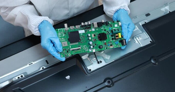 Technician wearing blue gloves holds a green circuit board while repairing the inside of a flat screen television.