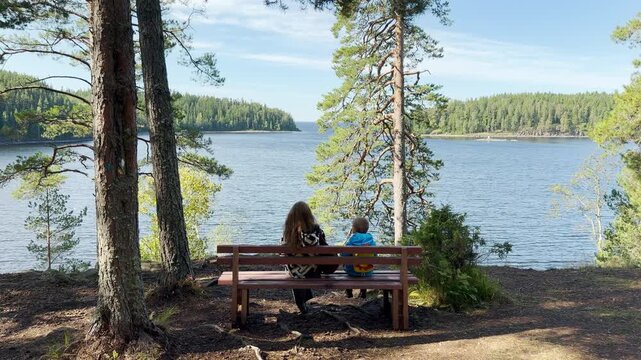 A woman and child sit peacefully on a bench overlooking the water on Valaam, Lake Ladoga. This serene moment evokes a warm, joyful, and tranquil feeling.