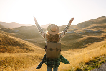 Fototapeta premium A female traveler with a backpack and a hat enjoys the mountain scenery and a sense of freedom at sunset. She walks along a path amidst the grass. Hiking and nature concept.