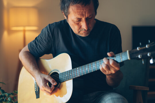 Man playing an acoustic guitar at home, close-up of hands strumming and fretting the strings, hobby and music concept in a cozy warm interior.