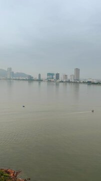 Aerial panoramic view of fishing boats sailing in the foreground with Quy Nhon city skyline in the background, Vietnam. Shot from Hai Minh peninsula featuring calm sea, coastal bay, and cityscape.