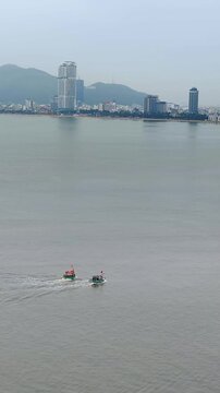 Aerial panoramic view of fishing boats sailing in the foreground with Quy Nhon city skyline in the background, Vietnam. Shot from Hai Minh peninsula featuring calm sea, coastal bay, and cityscape.
