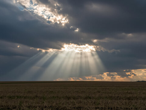 Dramatic sun rays breaking through dark storm clouds over a harvested field at sunset.