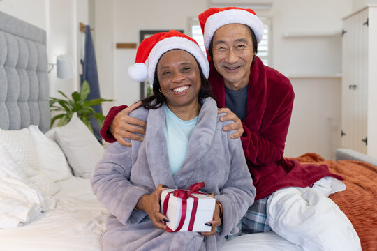 Senior Asian man and African woman sitting in bedroom wearing Santa hats holding gift box christmas