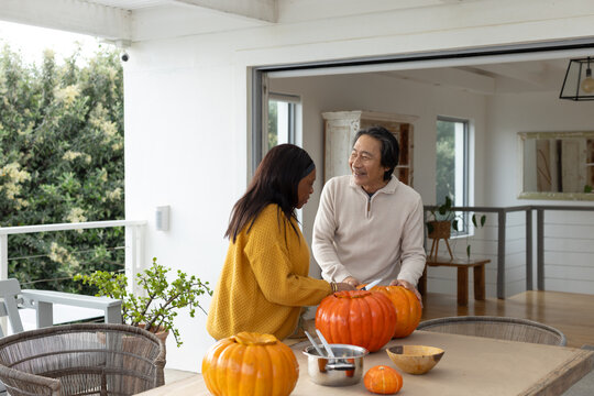 Senior Asian man, African American woman carving pumpkins on porch table with bowl for halloween