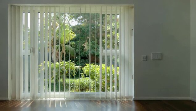 Relaxing 4K residential stock clip presenting white vertical window blinds adjusted halfway open revealing lush backyard trees outside while casting geometric linear shadows across a bright indoor