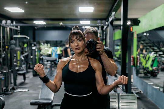 Woman flexing arms in gym mirror with photographer
