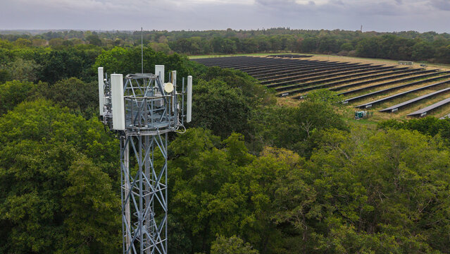 Fleet, England, United Kingdom - 18 September 2026: Aerial view of a mobile phone mast standing amidst lush green trees with a large solar panel farm visible in the background.