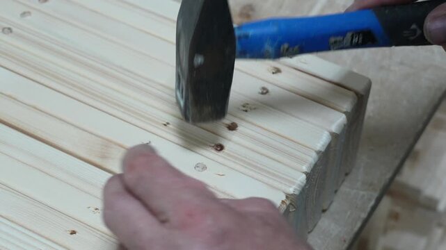 A craftsman uses a hammer to drive wooden dowels into pre-drilled holes in wooden slats during woodworking, joinery, and furniture making process