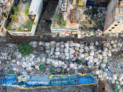 Dhaka, Bangladesh - 27 August 2026: Aerial view of a waste sorting area with large white bags of plastic recyclables, a blue tarp structure, and dense urban buildings.