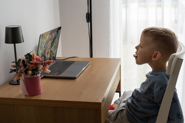 Young boy practices a tongue exercise during an online speech therapy session on a laptop at home. Concept of remote speech development, articulation training and accessible support for children. © Maryia