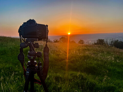 A close-up silhouetted view of a DSLR mounted on a tripod, positioned in a grassy field on a hilltop facing a vibrant sunset with lens flare streaming across the rural landscape.