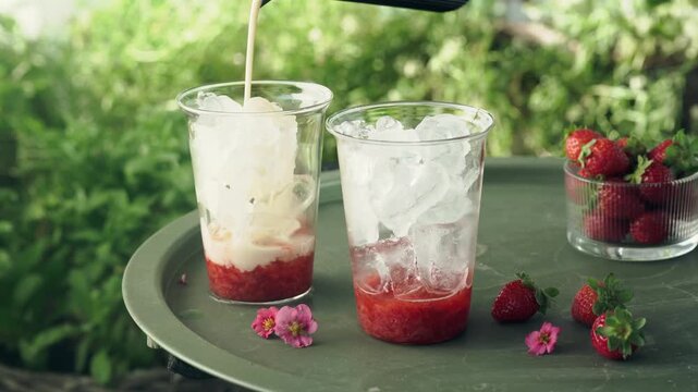 Milk is poured over fresh strawberry puree and ice to create a refreshing layered summer drink. Close-up drink preparation on an outdoor table surrounded by fresh strawberries and greenery