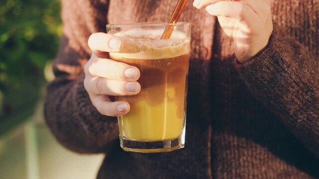Woman holding a glass of homemade pumpkin spice latte in warm autumn sunlight. Cozy seasonal lifestyle footage with a knitted brown cardigan